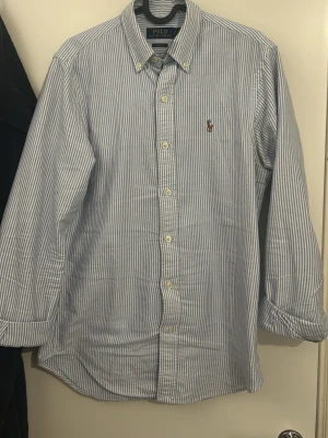 Randig skjorta från Polo Ralph Lauren - Klassisk blå- och vitrandig skjorta från Polo Ralph Lauren med button-down krage och broderad logga på bröstet. Skjortan har lång ärm och knäppning framtill. Perfekt för en stilren och avslappnad look. Lätt den fetaste skjortan till sommaren och är köpt ny men har växt ur den, nypris på denna är 2000 kr säljer för 549 kr och priset går att diskutera 🤩