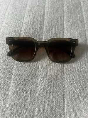 Chimi 04 mörkt olivgröna - Snygga solglasögon med fyrkantig form och transparent brun båge. Glasen är mörktonade och bågarna har två små silverprickar på varje sida framtill. Nyskick!