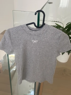 Grå t-shirt med rosettdetalj - En stilren grå t-shirt i bomull med korta ärmar och en liten vit broderad rosett framtill. Perfekt för en enkel och clean look. T-shirten har en klassisk passform och rund halsringning.