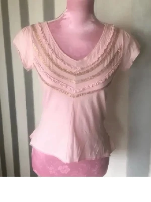 Ljusrosa blus med volang och spets - Söt ljusrosa blus med korta ärmar och v-ringad hals. Framsidan har dekorativa spetsdetaljer och volanger i flera lager. Nederkanten har en volang som ger ett extra feminint touch. Perfekt för dig som gillar romantiska och trendiga plagg.