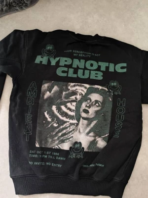 Svart sweatshirt Hypnotic Club - Cool svart sweatshirt från Hypnotic Club med grönt tryck på bröstet och stort grafiskt print på ryggen. Tröjan har rund hals, ribbade muddar och är tillverkad i mjukt bomullsmaterial. Perfekt för dig som gillar unika prints och streetwear.