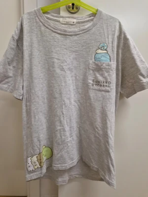 Grå Sumikko Gurashi t-shirt med ficka M - Grå t-shirt med tryck av Sumikko Gurashi-karaktärer på bröstfickan och nedre kanten. T-shirten har rund hals och korta ärmar, tillverkad i bomull. Extremt liten I storlek. Känns som XS eller mindre S.
