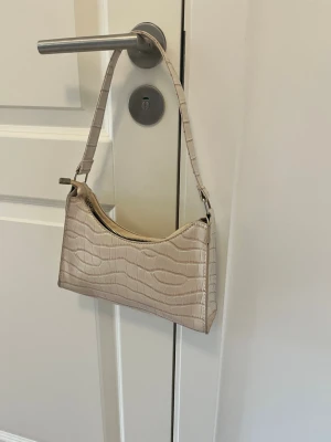 Beige axelremsväska i croco-mönster - Stilren beige handväska med axelrem och croco-mönstrad yta. Väskan har en rektangulär form och stängs med dragkedja upptill. Perfekt för dig som gillar trendiga accessoarer med en clean look. Materialet är konstläder.