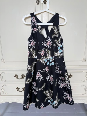Svart blommig klänning - Supersnygg svart klänning med blommigt mönster i rosa, gult och blått från Oasis. Klänningen har en kort längd, omlott-detalj framtill och kors i ryggen och är lite puffig nertill. Perfekt för dig som gillar en feminin och trendig stil. Materialet känns mjukt och följsamt. Är en liten felsömn på vänster bröst men syns knappt. 