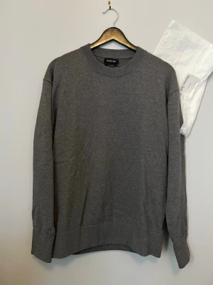 Grå stickad tröja från Massimo Dutti - Snygg grå stickad tröja från Massimo Dutti i 100% bomull. Tröjan har rund halsringning, långa ärmar och ribbade muddar vid hals, ärmslut och nederkant. Perfekt för dig som gillar stilrena och klassiska plagg.