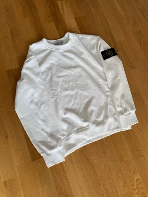 Vit sweatshirt från Stone Island - Vit sweatshirt från Stone Island med klassisk rund hals och ribbade muddar. Tröjan har den ikoniska svarta logotyp-patchen med gult broderi på vänster ärm. Perfekt för en clean och stilren look.