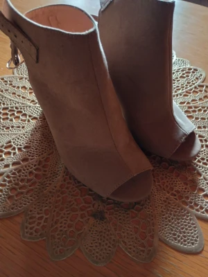 Beige peep toe stövletter i mocka - Snygga beige stövletter med peep toe och öppen häl. Skorna har en trendig siluett med spänne runt ankeln och är tillverkade i mjuk mocka. Perfekta för dig som vill ha en stilren och modern look.