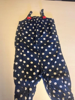 Blå prickig jumpsuit med rosetter - Supersöt blå jumpsuit med vita stora prickar och röda rosetter på axelbanden. Smock upptill och resår vid bensluten ger en skön passform. Perfekt för dig som gillar lekfulla och trendiga plagg. Materialet känns lätt och luftigt.