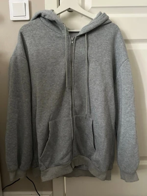 Grå overzised zip hoodie med huva - En klassisk grå oversized zip hoodie med dragkedja framtill och två stora fickor. Hoodien har justerbar dragsko i huvan och ribbade muddar vid ärmar och nederkant. Perfekt för en avslappnad och chill stil. Materialet är mjukt och bekvämt, troligtvis bomullsblandning.