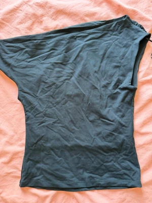 Svart oneshoulder topp från Gina Tricot - Trendig svart oneshoulder topp i mjukt material. Snygg asymmetrisk design med en ärm och en bar axel, perfekt för dig som vill sticka ut lite extra. Passar till både jeans och kjol för en cool look. Lite nopprig på axeln