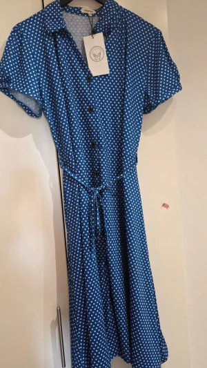 Blå prickig klänning med knytband - Snygg blå klänning med vita prickar, knappar framtill och knytband i midjan. Kortärmad modell med krage och lätt utställd kjol. Perfekt för en fräsch och somrig look.