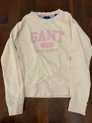 Beige sweatshirt från GANT - Snygg beige sweatshirt från GANT med rosa tryck framtill där det står 'GANT 1949 NEW HAVEN'. Tröjan har rund halsringning, långa ärmar och ribbade muddar. 