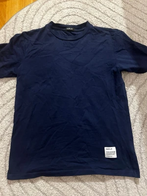 Mörkblå t-shirt från Replay - Snygg mörkblå t-shirt från Replay med klassisk passform och rund hals. T-shirten har korta ärmar och en liten vit patch med logga nertill samt tryck på ryggen vid axeln. Perfekt till jeans eller shorts.