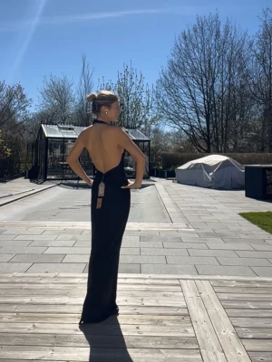 Svart långklänning med öppen rygg - Elegant svart långklänning med halterneck och helt öppen rygg. Klänningen har en figurnära passform och faller snyggt längs kroppen. Perfekt för dig som vill ha en stilren och sofistikerad look. Endast använd en gång. Helt ny