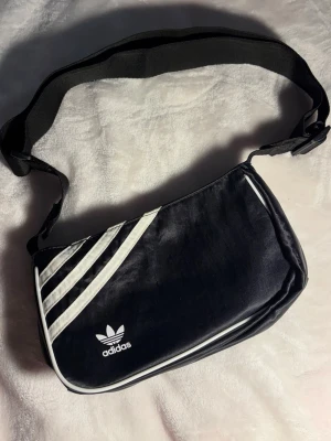 Svart axelväska från Adidas - Snygg svart axelväska från Adidas med klassiska vita ränder och Adidas-logga på framsidan. Väskan har justerbar axelrem och dragkedja upptill. Tillverkad i ett glansigt tygmaterial som ger en sportig vibe. Perfekt för dig som gillar streetwear och vill ha en praktisk väska.