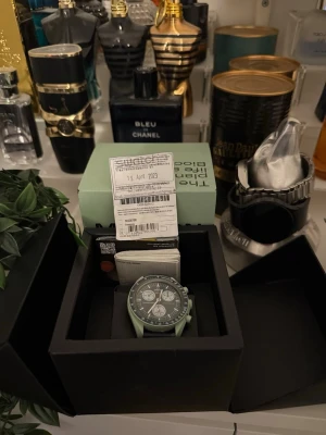 Swatch x Omega MoonSwatch klocka - Säljer en Swatch x Omega MoonSwatch med svart urtavla och ljusgrön boett. Klockan har kronograf och svart armband. Kommer i originalbox med tillbehör. Perfekt för dig som gillar coola och unika accessoarer. Kan tänka mig byta den mot en annan klocka, parfymer eller annat.