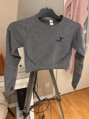 Grå cropped träningströja med tumhål - Snygg grå långärmad cropped träningströja med svart logga på bröstet. Tröjan har tumhål i ärmsluten och är tillverkad i ett stretchigt funktionsmaterial som passar perfekt till träning.