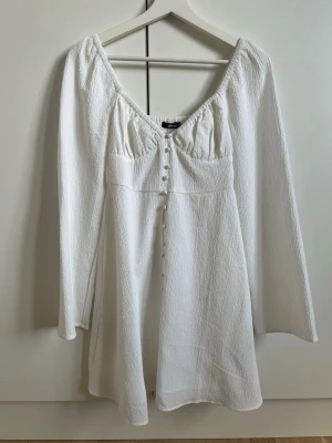 Vit kortklänning från Gina Tricot - Superfin vit kortklänning från Gina Tricot med långa ärmar och knappar framtill. Klänningen har en lätt rynkad byst och v-ringad halsringning. Tillverkad i ett strukturerat tyg som ger en snygg och trendig look. Användes halva dagen på min student, så är i väldigt fint skick!☺️