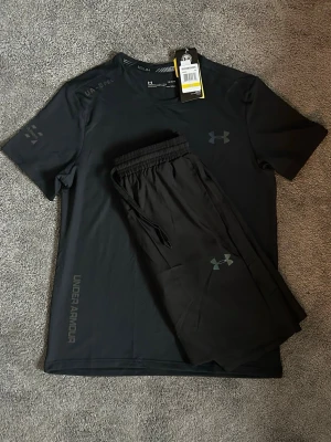 Svart träningsset från Under Armour - Snyggt svart träningsset från Under Armour med t-shirt och shorts. T-shirten har diskret logga på bröstet och tryck på ärmen, medan shortsen har elastisk midja med dragsko och logga på benet. Perfekt för gymmet eller löprundan.
