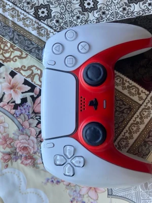 Vit och röd PlayStation 5 DualSense - PlayStation 5 DualSense handkontroll i vit och röd färg med klassisk design och svarta analoga spakar. Kontrollen har touchpad, ikoniska PlayStation-knappar och är tillverkad i plast. Perfekt för gaming på PS5.