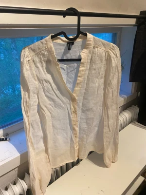 Krämvit blus från Vero Moda - Stilren krämvit blus från Vero Moda med v-ringning och knappar framtill. Blusen har långa ärmar och är tillverkad i ett lätt, luftigt tyg som ger en avslappnad känsla. Perfekt att matcha med jeans eller kjol för en trendig look. är inte lika genomskinlig i verkligheten 