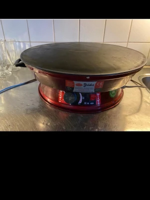 Röd elektrisk crêpemaker Yildiz - Elektrisk crêpemaker från Yildiz i röd metall med stor, rund svart stekyta. Perfekt för att göra pannkakor eller crêpes hemma. Utrustad med vred för temperaturkontroll och tydliga indikatorlampor. Snygg och robust design som passar på köksbänken.