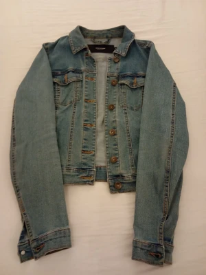 Ljusblå jeansjacka från Vero Moda - Trendig ljusblå jeansjacka från Vero Moda med klassisk krage, bröstfickor med lock och knappar framtill. Jackan har en rak, något kortare passform och detaljerade sömmar. Perfekt för en avslappnad streetstyle-look.