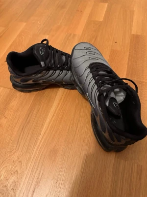 Nike TNS - Snygga sneakers i grått och svart med vågformade detaljer och svarta snören. Skorna har en sportig look med mesh-liknande material och svart sula. Perfekta för dig som gillar en modern och stilren stil.