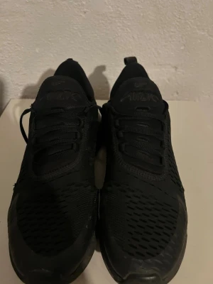 Svarta Nike Air Max 270 sneakers - Säljer ett par helsvarta Nike Air Max 270 sneakers med klassisk luftig mesh-ovandel och synlig Air-enhet i hälen. Skorna har en sportig design med rundad tå, svarta skosnören och diskreta loggor på plösen och sidan. Perfekta för en streetwear-look.