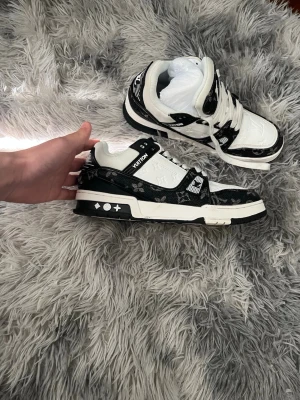 Louis Vuitton svartvita sneakers - Säljer ett par snygga Louis Vuitton sneakers i svart och vitt med klassiskt monogrammönster. Skorna har lågt skaft, vita snören och detaljer i svart läder samt präglad logga på sidan. Perfekta för dig som vill sticka ut med exklusiv streetstyle.