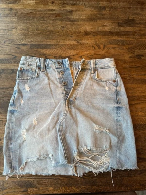 Ljusblå jeanskjol med slitningar - Trendig ljusblå jeanskjol med råa kanter och slitna detaljer framtill. Kjolen har knappar i gylfen, bälteshällor och klassiska jeansfickor. Perfekt för dig som gillar en avslappnad och edgy stil.