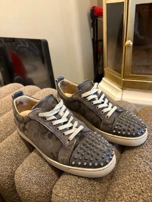 Unika Christian Louboutins - Snygga gråa sneakers från Christian Louboutin med ikoniska röda sulor och coola silverfärgade nitar på tån. Skorna har vita snören och är tillverkade i mocka med diskret mönster. Perfekta för dig som vill sticka ut med din stil. Skick 7/10