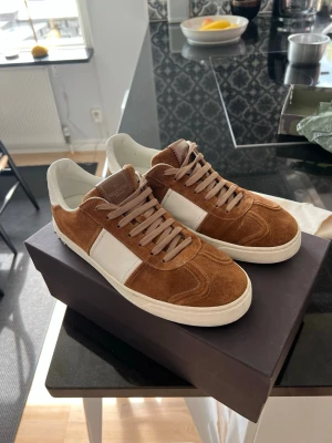 Bruna och vita sneakers från Valentino - Snygga sneakers från Valentino i brunt mocka och vitt läder. Skorna har vita detaljer på sidan och hälen samt coola nitar bak på sulan. Klassisk låg modell med bruna skosnören och vit sula. Perfekta för dig som vill ha en stilren men ändå unik look.