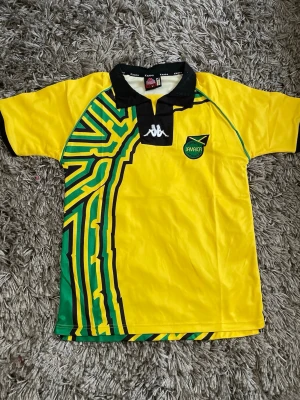 Jamaica fotbollströja från Kappa - Gul Jamaica fotbollströja från Kappa med svart krage och gröna samt svarta detaljer. Tryckt Kappa-logga på bröstet och Jamaica Football Federation-märke. Unik grafisk design i grönt, gult och svart på ena sidan. Perfekt för dig som gillar fotboll och vill sticka ut.