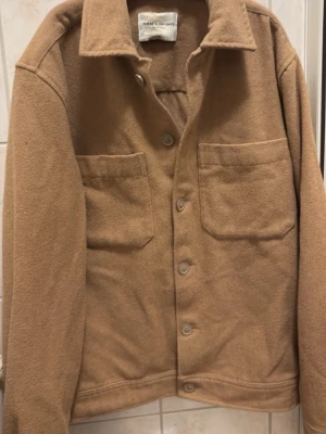 Beige overshirt från The Everyday Café - Stilren beige overshirt i mjukt material från The Everyday Café. Skjortjackan har två stora bröstfickor, knäppning framtill och klassisk krage. Perfekt för lager-på-lager och enkel att matcha med olika outfits.