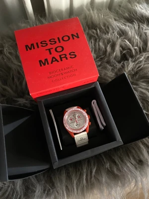 Omega x Swatch Mission to Mars klocka - Säljer en Omega x Swatch Speedmaster MoonSwatch Mission to Mars. Klockan har en vit urtavla, röd boett och vita detaljer. Biokeramisk konstruktion och textilarmband med Omega-logga. Kommer i originalförpackning.