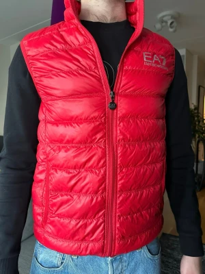 Armani dunväst - Röd dunväst från Emporio Armani EA7 med quiltad design och dragkedja framtill. Västen har två sidofickor och logga på bröstet. Materialet är glansigt och lätt, vilket gör den väldigt bekväm! Storlek M💫