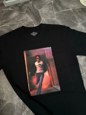 Djo Joe keery Tshirt Steve harrington - Storlek S, lite oversize. Svart t-shirt med stort färgstarkt tryck framtill som visar en person som springer ut från en byggnad. Klassisk rund hals och korta ärmar. Materialet är mjuk bomull som känns skön mot huden. Perfekt för dig som gillar streetwear och unika prints.