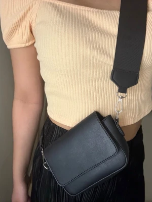 Svart axelväska med bred rem - En oanvänd stilren svart axelväska med bred justerbar rem och silverfärgade metalldetaljer. Väskan har en minimalistisk design med slät yta och är tillverkad i syntetmaterial. Perfekt för att bära det viktigaste och ger en cool touch till din outfit. 