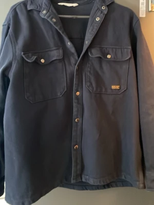 Mörkblå overshirt från H&M - Snygg mörkblå overshirt från H&M med två stora bröstfickor med lock och tryckknappar. Overshirten har klassisk krage och raka ärmar, perfekt att bära som lager. Materialet är kraftigt och ger en cool look.