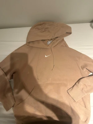 Beige hoodie från Nike med huva - Säljer en beige hoodie från Nike med klassisk vit swoosh på bröstet. Hoodien har en stor huva med dragsnören och en praktisk magficka. Materialet är mjukt och bekvämt, perfekt för chill dagar. Ribbade muddar på ärmar och nederkant.