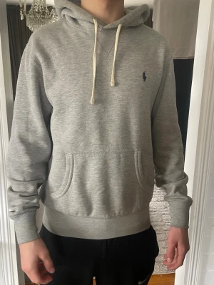  Grå ralph lauren hoodie med huva - Säljer en stilren grå hoodie med huva och dragsnören. Hoodien har en klassisk passform, ribbade muddar vid ärmslut och nederkant samt fickor på sidorna. Perfekt för en avslappnad och bekväm look. Materialet känns mjukt och skönt mot huden.