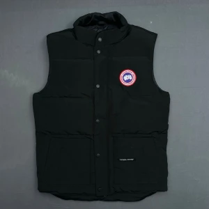 Svart dunväst från Canada Goose - Snygg svart dunväst från Canada Goose med klassisk logga på bröstet. Västen har hög krage, tryckknappar framtill och två stora fickor. Perfekt för lager-på-lager och håller dig varm under kyliga dagar. Jag skulle säga att den passar som en stor M eller liten L