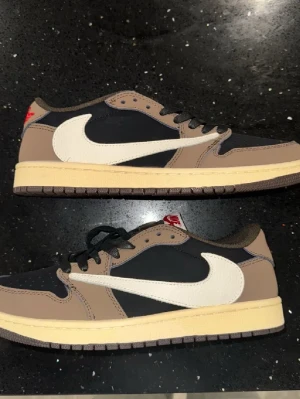 Nike Air Jordan 1 Low Travis Scott - Säljer ett par Nike Air Jordan 1 Low Travis Scott sneakers med bruna och svarta paneler, vit omvänd swoosh och beige mellansula. Skorna har klassisk låg profil, svarta skosnören och röda detaljer på hälen. Perfekt för dig som vill sticka ut med en ikonisk modell.