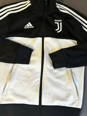 Juventus Adidas svartvit träningsjacka - Juventus träningsjacka från Adidas i svart och vitt med dragkedja och ståkrage. Jackan har två sidofickor och klassiska Adidas-ränder på axlarna. Materialet är polyester och logotypen är broderad på bröstet. Perfekt för fotboll och sport.