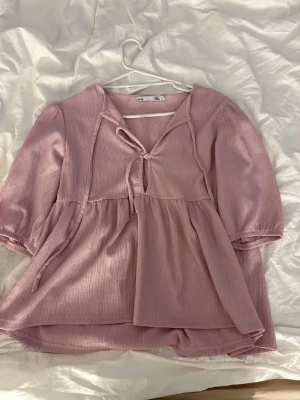 Ljusrosa blus med knyt och puffärm - Söt ljusrosa blus med knyt framtill och v-ringning. Blusen har puffiga trekvartsärmar och ett lätt, luftigt tyg med struktur. Perfekt för en trendig och avslappnad look.