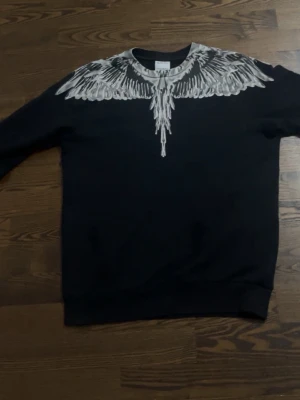 Marcelo burlon tröja äkta - Säljer en svart sweatshirt från Marcelo Burlon med ikoniskt vitt vingeprint runt halsen och axlarna. Tröjan har rund halsringning och ribbade muddar vid ärmslut och nederkant. Perfekt för dig som vill sticka ut med en unik design.