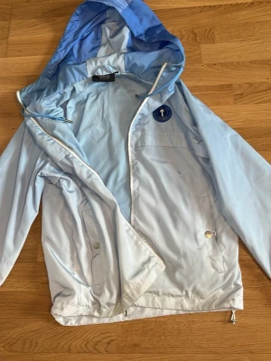 Ljusblå vindjacka från Trapstar - Snygg ljusblå vindjacka från Trapstar med huva och dragkedja. Jackan har två fickor med tryckknappar, justerbar nederkant och cool Trapstar-logga i blått på ryggen. Materialet är tunt och lätt, perfekt för blåsiga dagar.