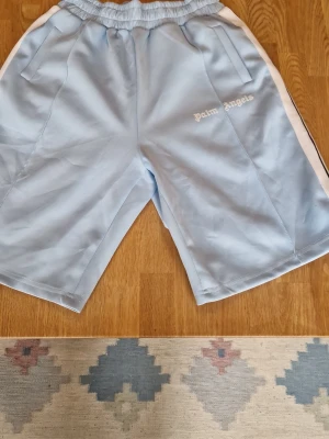 Ljusblå Palm Angels shorts - Säljer ett par ljusblå shorts från Palm Angels med vita sidoränder och broderad logga på vänster ben. Shortsen har elastisk midja och två fickor fram. Perfekta för en avslappnad streetwear-look.