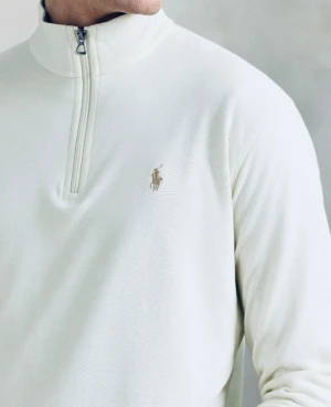 Vit half-zip tröja från Polo Ralph Lauren - Klassisk vit långärmad tröja från Polo Ralph Lauren med half-zip och broderad logga på bröstet. Tröjan har hög krage och ribbade muddar vid ärmslut och nederkant. Perfekt för en clean och stilren look.